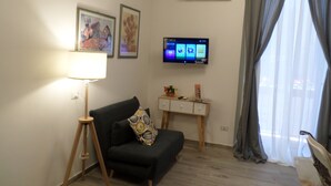 Un salón con un sofá negro, una lámpara de pie, un televisor de pantalla plana y una mesita con adornos.
