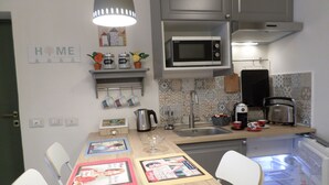 Una cocina funcional con microondas, hervidor, tostadora y refrigerador. Hay sillas blancas alrededor de una pequeña mesa de comedor con posavasos.