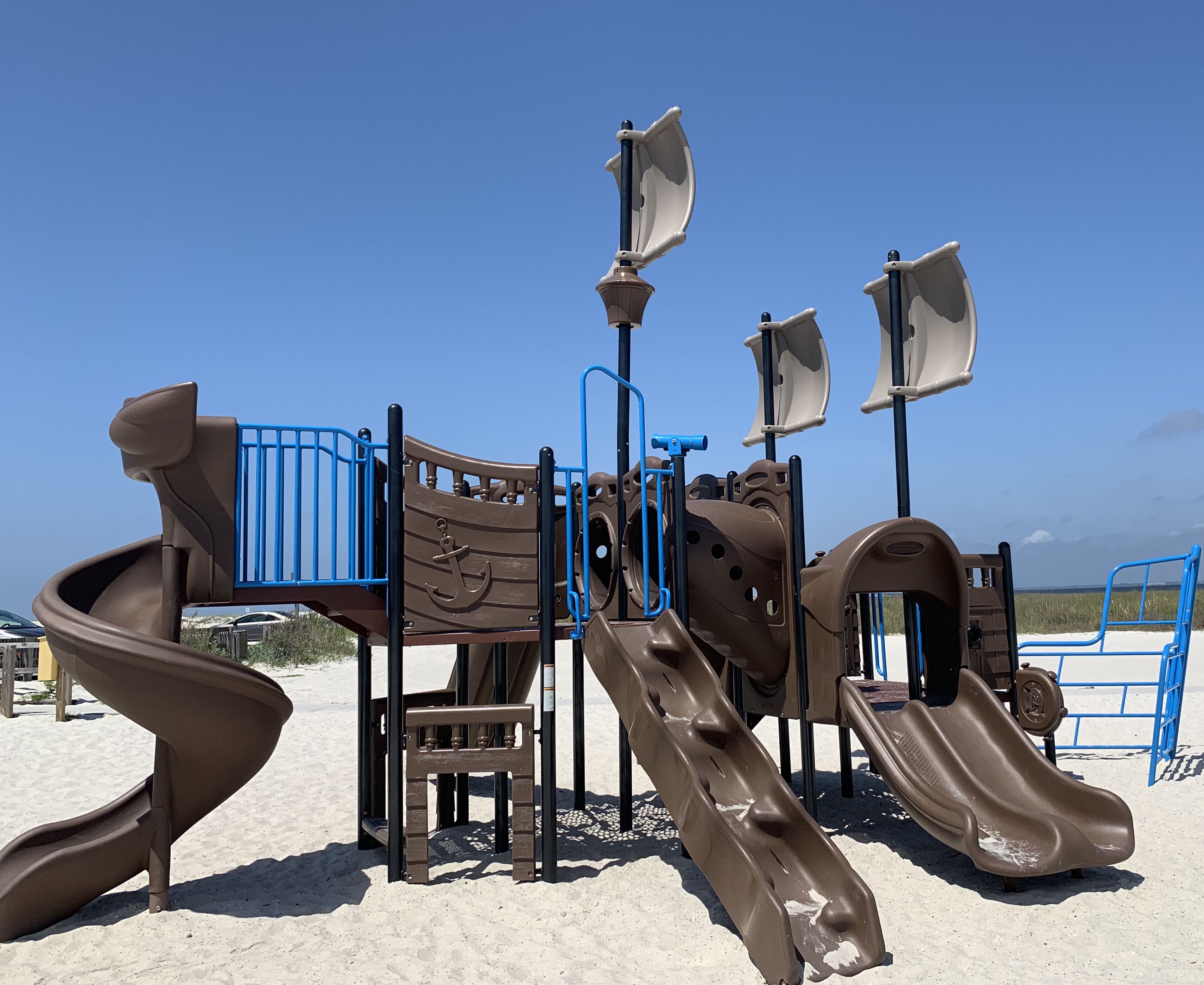 Ein Spielplatz mit Rutschen und Klettergerüsten auf sandigem Untergrund, im Hintergrund ein klarer Himmel.