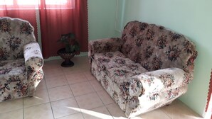 Una habitación con un sofá y un sillón estampado con motivos florales, una planta en maceta y una ventana con cortinas rojas.