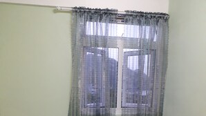 Una cama individual con estructura de madera y ropa de cama gris en una habitación con paredes verde claro y una ventana con cortinas translúcidas.