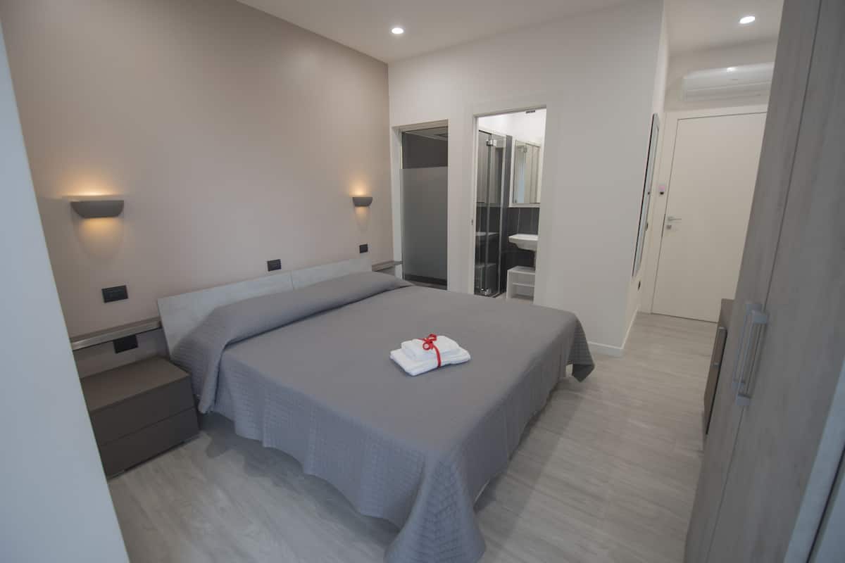 Habitación de hotel moderna con una cama grande, una mesita de noche, un armario y un baño visible a través de una puerta de cristal.