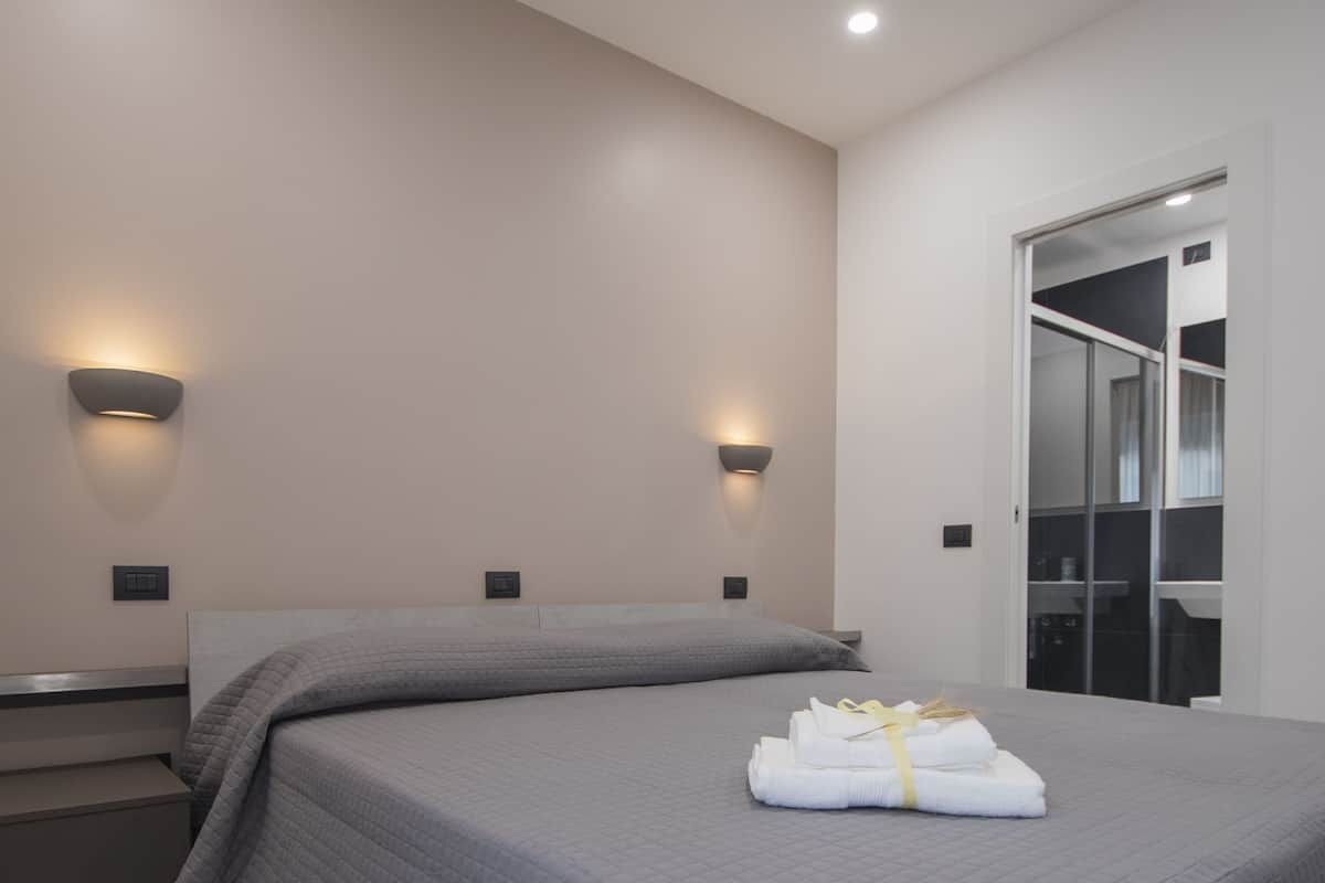 Una habitación de hotel moderna con una cama grande, dos lámparas adosadas a la pared, un armario y un baño visible a través de una puerta abierta.