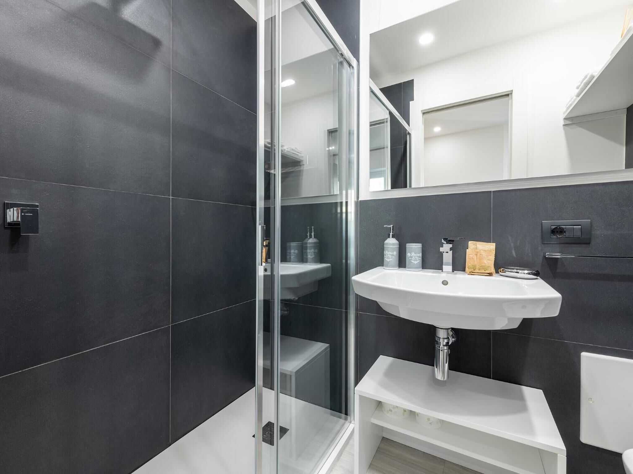 A modern bathroom with dark tiles, a glass shower enclosure, a white sink, and a mirror.