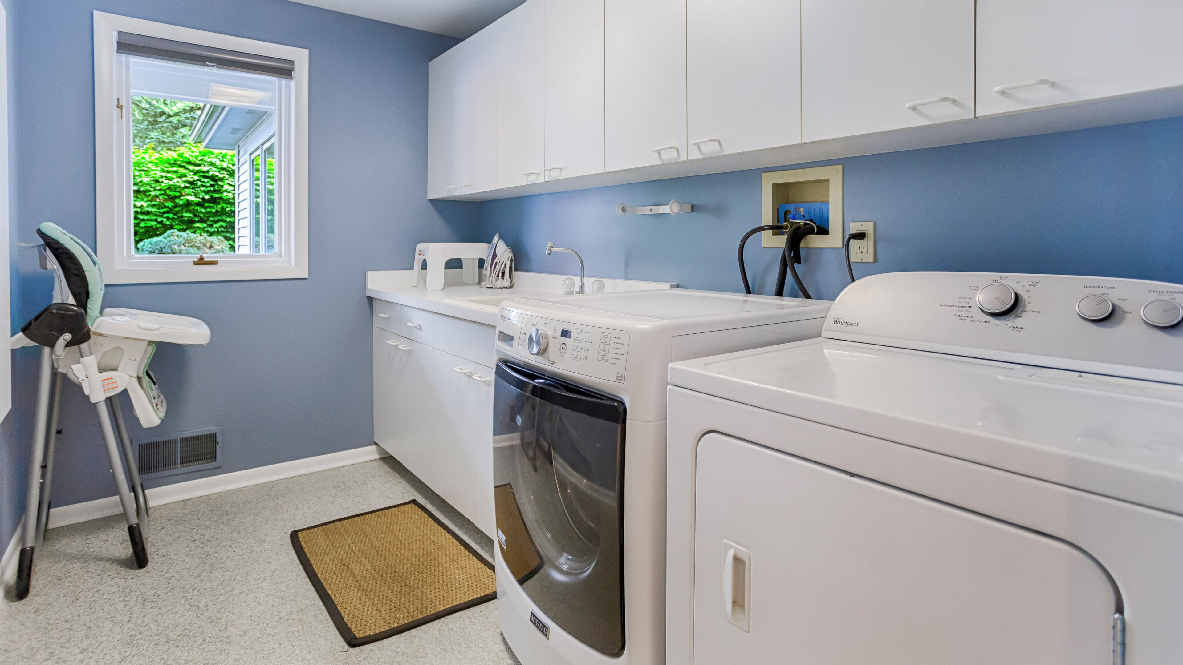 A laundry room with a front-loading washer and dryer, a small window with a view of greenery, and a white countertop with a microwave and other appliances.