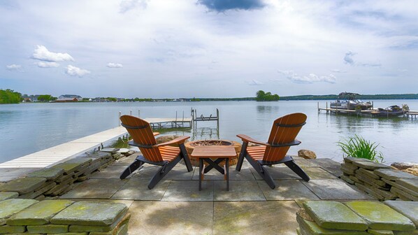 A lakeside patio with two Adirondack chairs and a small table, overlooking a calm lake with docks and boats.
