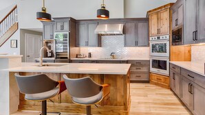 Une cuisine moderne dotée d’un îlot central, de deux tabourets de bar, d’un réfrigérateur en acier inoxydable, de deux fours et de meubles de rangement en bois.