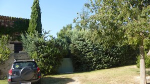 A gravel driveway leading to a parked SUV in a residential area with a stone wall, a wooden gate, and dense greenery. - rental house in the Luberon (ROUSSILLON)