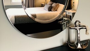Un miroir rond reflète un lit soigneusement fait, avec des oreillers, deux lampes de chevet et une table de chevet sur laquelle se trouve une lampe.