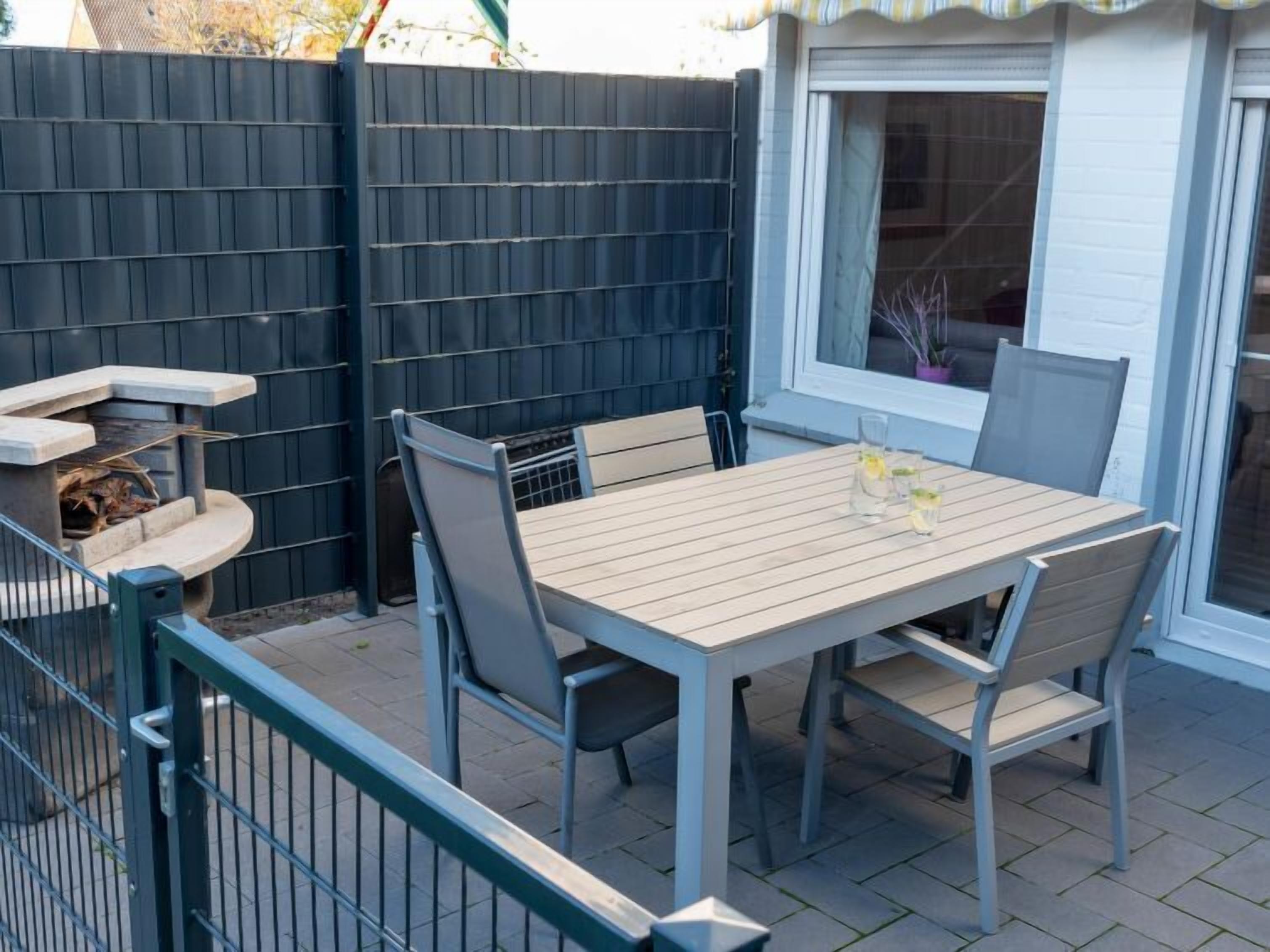 En uteplats med ett träbord och stolar, en liten grill och ett fönster med utsikt över huset.