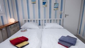 Cama doble con ropa de cama blanca, dos almohadas y ropa doblada a cada lado. Hay una mesita de noche con una lámpara y una pequeña mesa con un libro. La habitación tiene papel tapiz a rayas y una puerta blanca.
