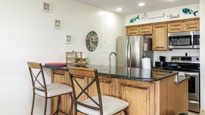 A kitchen with wooden cabinets, a stainless steel refrigerator, a microwave, and a sink. There are two bar stools and a clock on the wall.