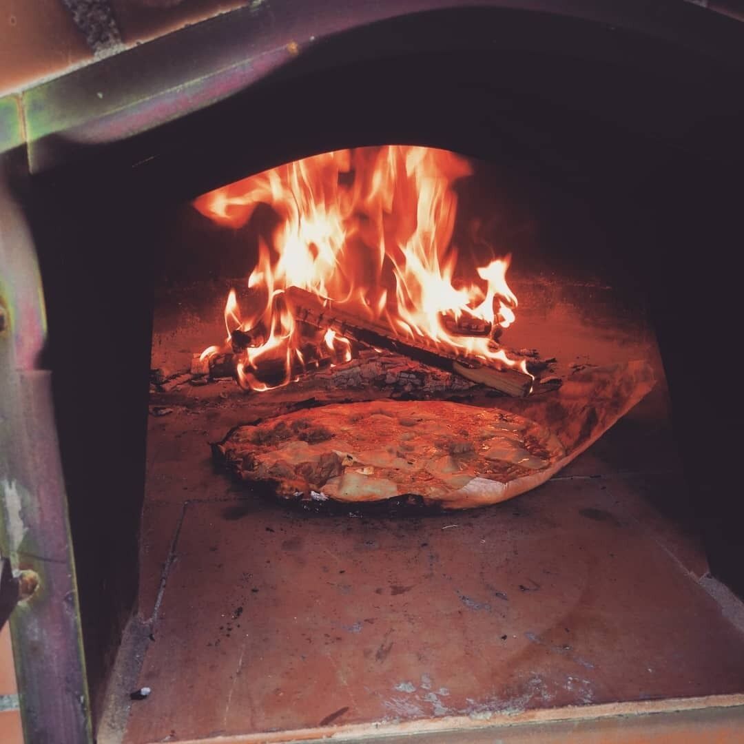 A wood-fired oven with a fire burning inside, logs stacked on the left, and a pizza stone in the center.