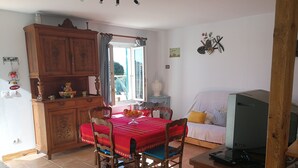 A dining area with a red tablecloth, wooden chairs, and a cabinet. There is a window with curtains, a television on a wooden stand, and a small table with a bowl of fruit.