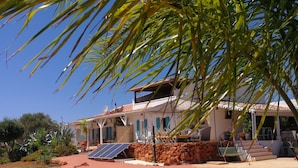 A clear blue pool surrounded by a stone patio, with a house in the background featuring a solar panel and a stone wall.