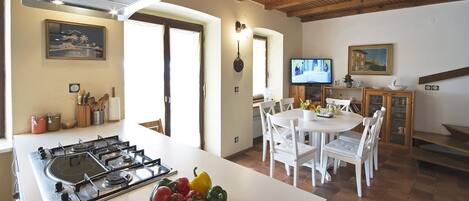 A kitchen with a gas stove, a bowl of tomatoes and peppers, a dining area with white chairs, and a television.