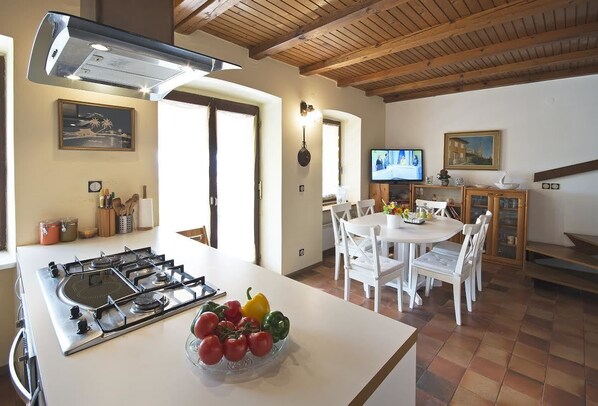 A kitchen with a gas stove, a bowl of tomatoes and peppers, a dining area with white chairs, and a television.