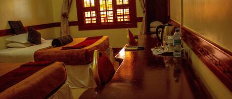 Habitación de hotel con dos camas individuales, una mesa de madera con una silla, una ventana con cortinas y una tetera sobre la mesa.