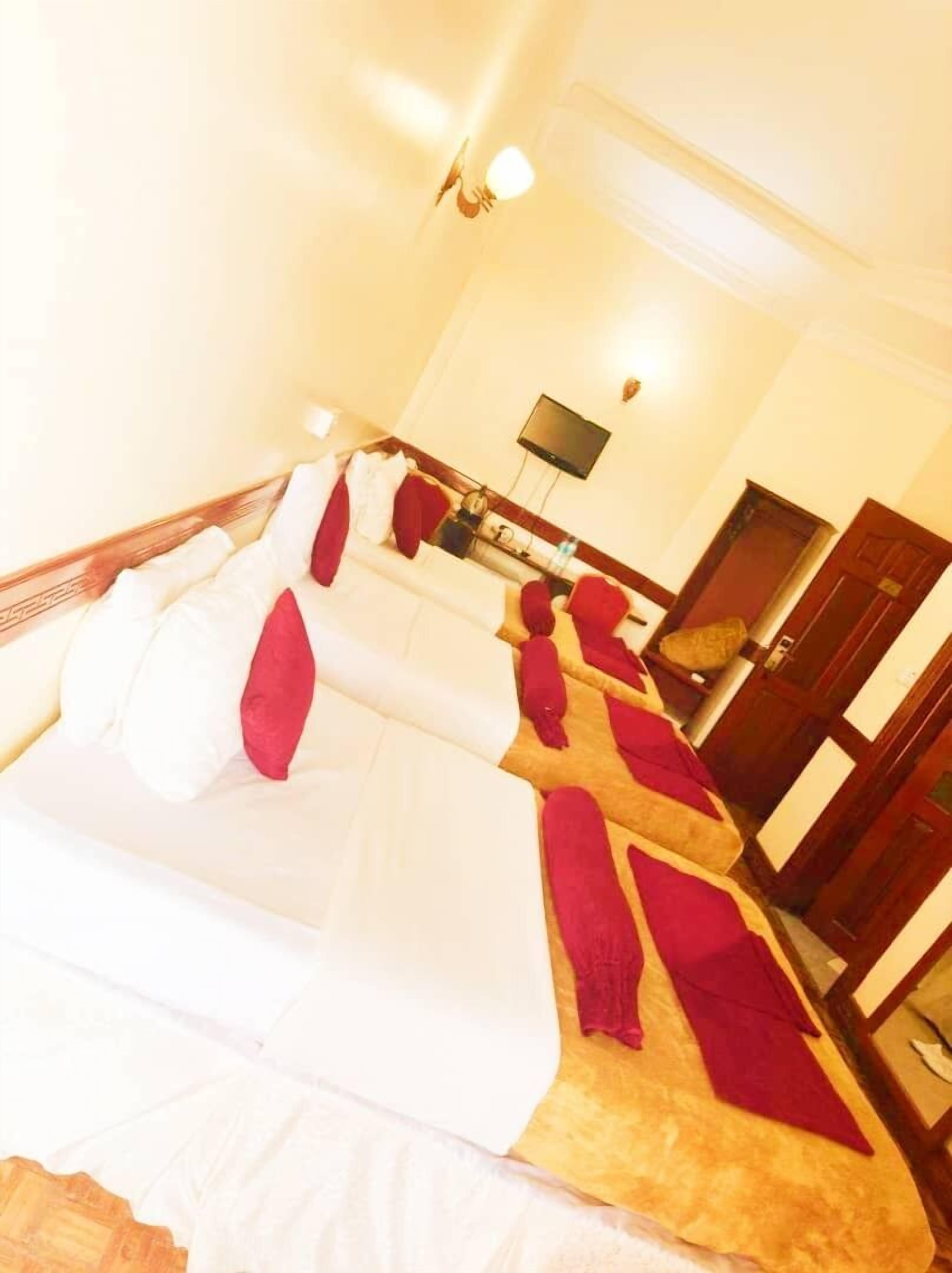 A hotel room with four beds arranged in a row, each with white bedding and red pillows. There is a flat-screen TV mounted on the wall above the beds, and a wooden door to the right.