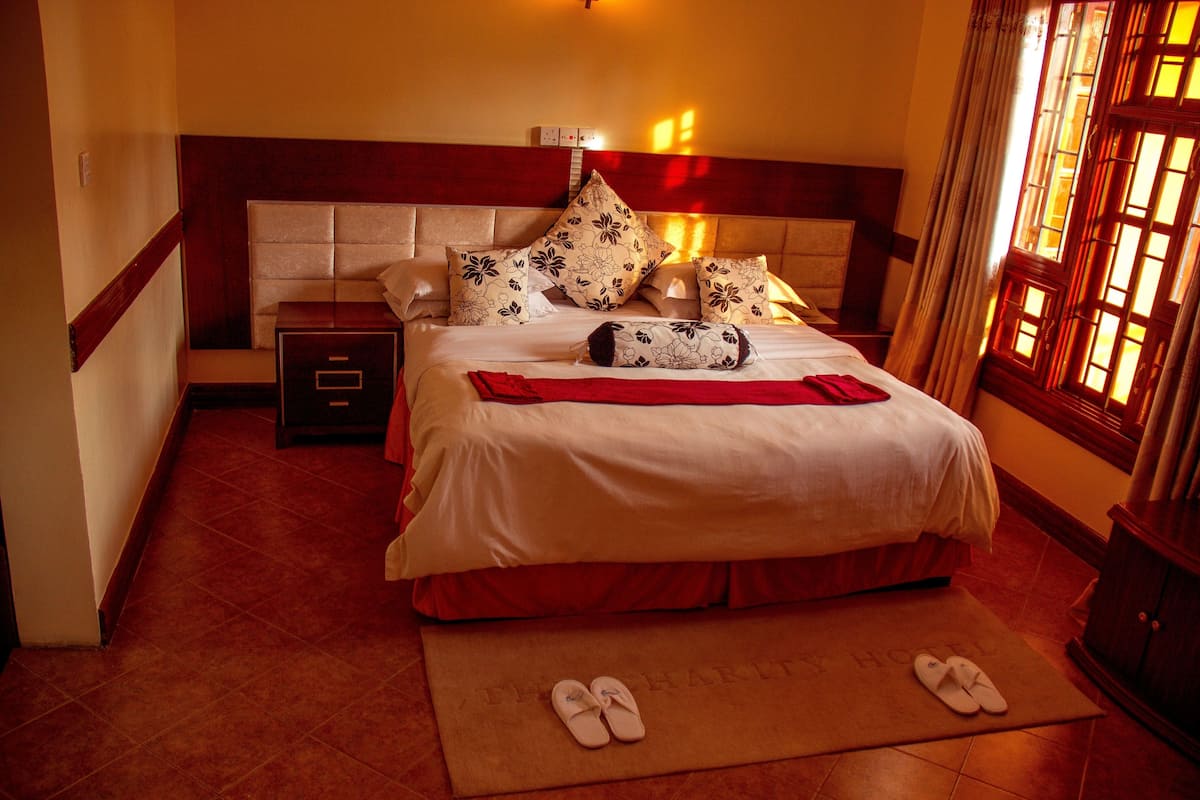 A hotel room with a bed, a nightstand, a red mat, and slippers.