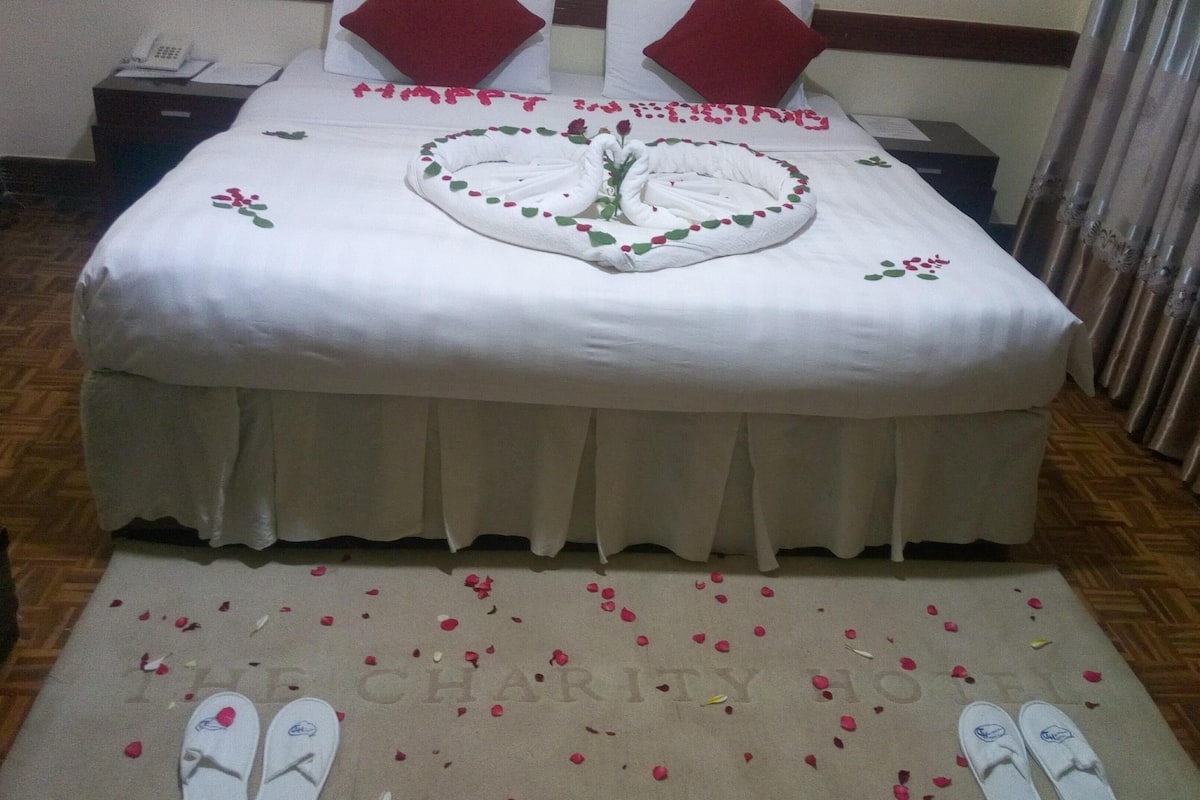 A hotel room with a neatly made bed, a heart-shaped towel arrangement, and a welcome mat with 'WELCOME' and 'CHARITY' inscribed.