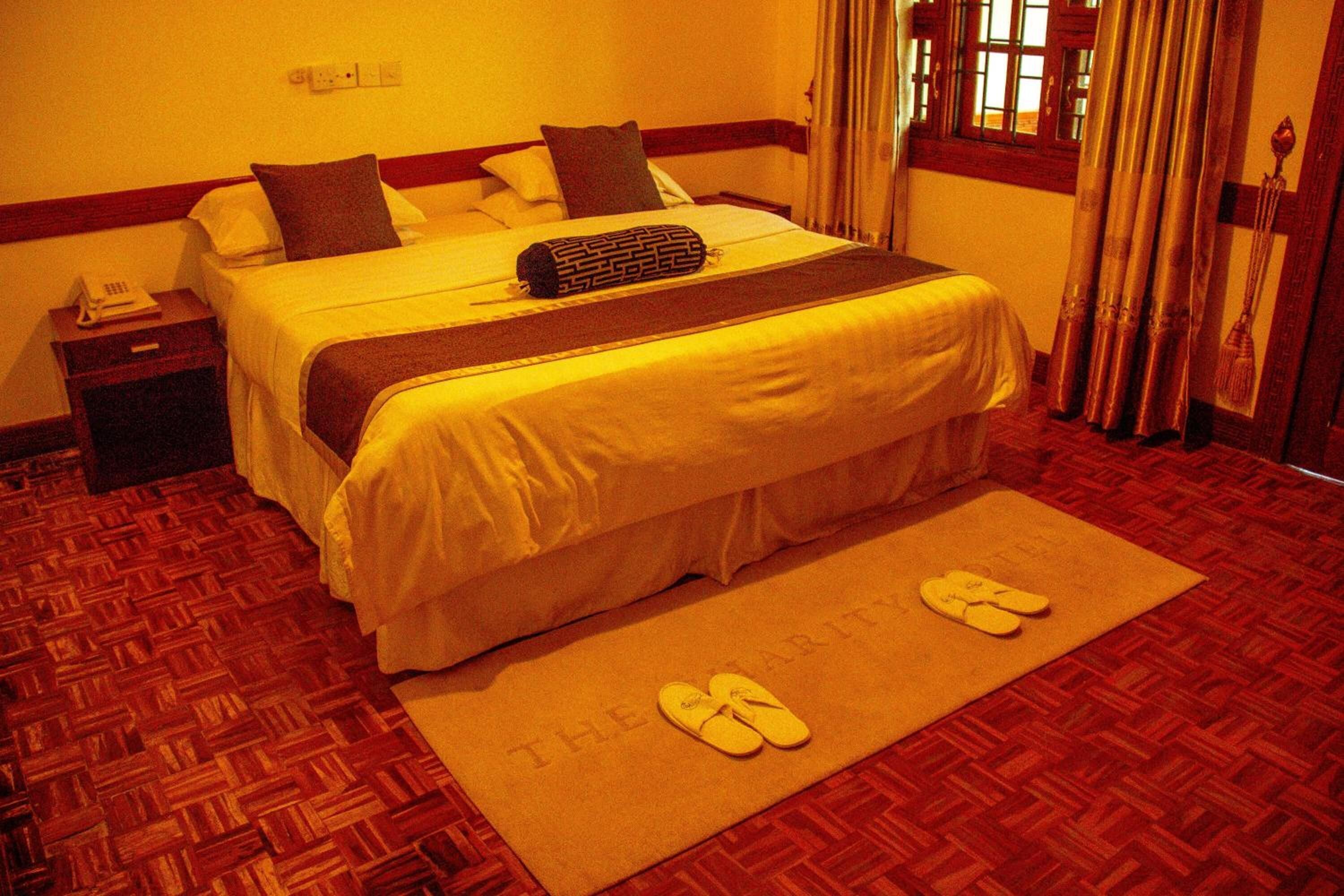 Quarto de hotel com cama, mesa de cabeceira, telefone, janela com cortinas e um par de chinelos sobre um tapete.