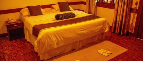 Quarto de hotel com cama, mesa de cabeceira, telefone, janela com cortinas e um par de chinelos sobre um tapete.