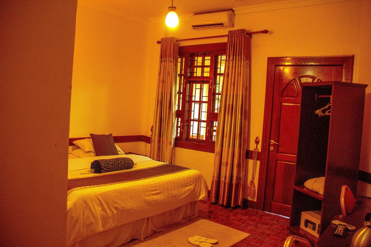 A hotel room with a bed, a wooden wardrobe, a mirror, and a window with curtains.