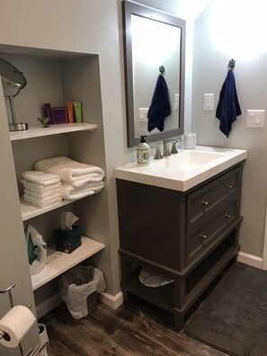 Casa de banho moderna com toucador escuro, prateleiras brancas com toalhas, espelho e suporte para papel higiénico.