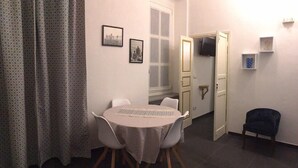 Ein Essbereich mit einem runden Tisch, Stühlen und einem Fenster. An der Wand hängen zwei gerahmte Bilder, ein Fernseher und eine Tür zu einem anderen Raum.