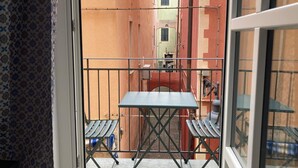 Ein Balkon mit Tisch und Stühlen, einem gemusterten Vorhang und Blick auf eine enge Straße mit Häusern.