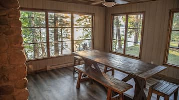 Un comedor rústico con una gran mesa de madera y bancos, ventanas amplias con vistas a árboles, y paredes de madera.