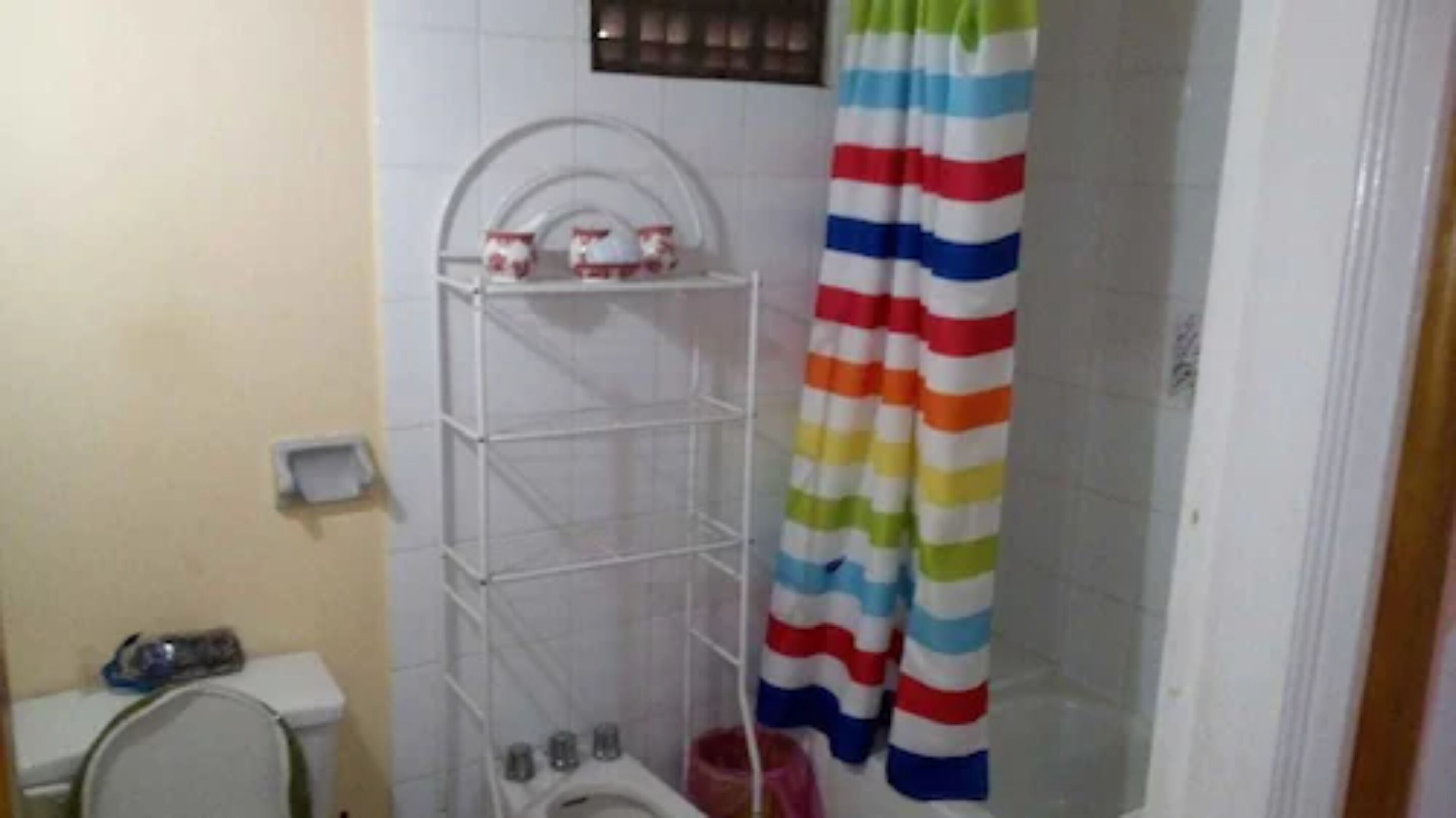 A small bathroom with a white tiled wall, a white toilet, a white shelf unit, and a striped shower curtain.