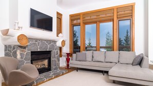 Una sala de estar con chimenea de piedra, un sofá seccional gris, un sillón moderno y un amplio ventanal con vista a árboles.
