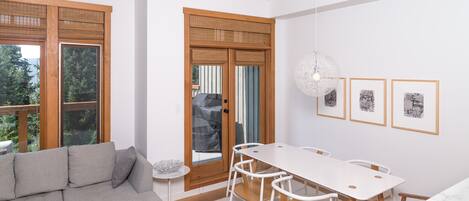 A modern living room with a grey sofa, a white dining table with chairs, and a large window with wooden shutters.