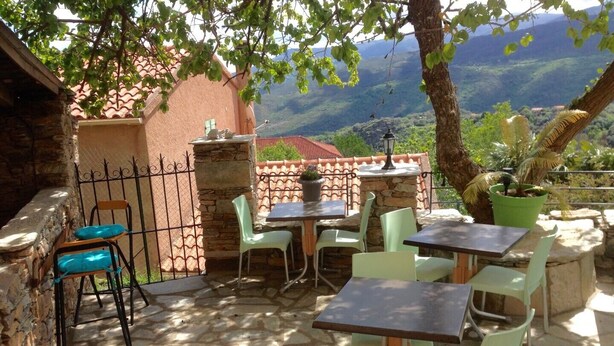 Un patio al aire libre con suelo de piedra, sillas y mesas verdes, rodeado por un muro de piedra y vistas a un paisaje montañoso.