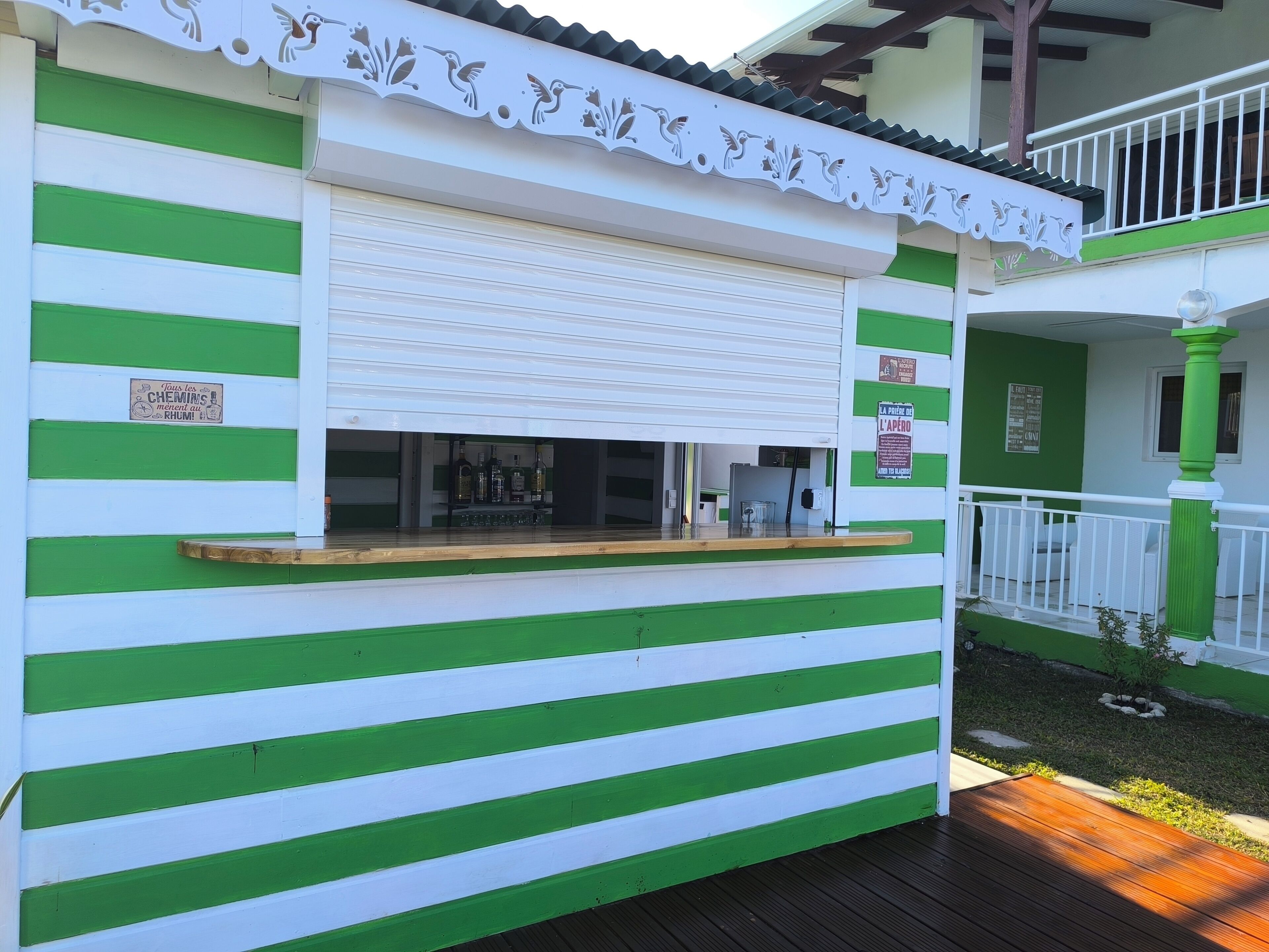 Un espace bar extérieur aux rayures vertes et blanches, doté d’un comptoir, d’un volet fermé et d’une enseigne avec du texte.