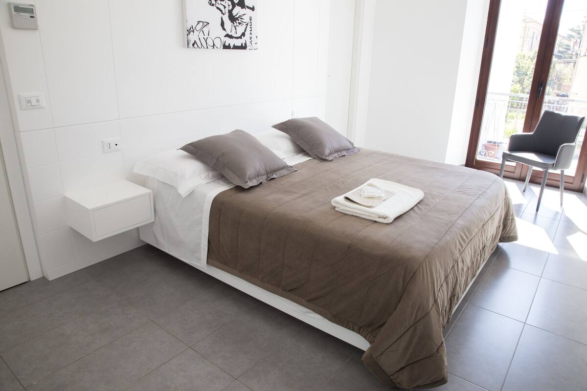 Un dormitorio moderno con una cama, dos almohadas, una mesita de noche y una silla junto a la ventana.