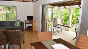 A room with a sofa, a TV, a dining table, and chairs. There is a sliding glass door leading to an outdoor area with a bench and greenery. - Rarotonga Island Villa 1 (Papaaroa)