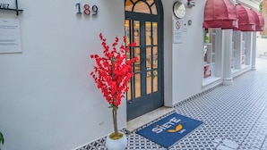 Entrada de hotel con puerta numerada, felpudo y planta en maceta.