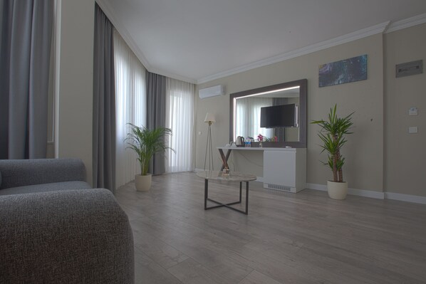 A modern hotel room with a grey sofa, a round coffee table, a flat-screen TV, and two potted plants. - Siete Hotel (Antalya)