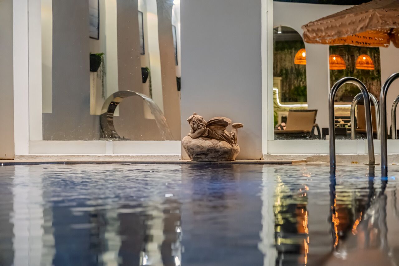 A swimming pool with a decorative sculpture, a glass wall reflecting interior lighting, and a glimpse of a dining area with tables and chairs.