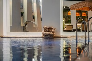 Una piscina con una escultura decorativa, un muro de cristal que refleja luces del interior y una vista de un comedor con mesas y sillas.