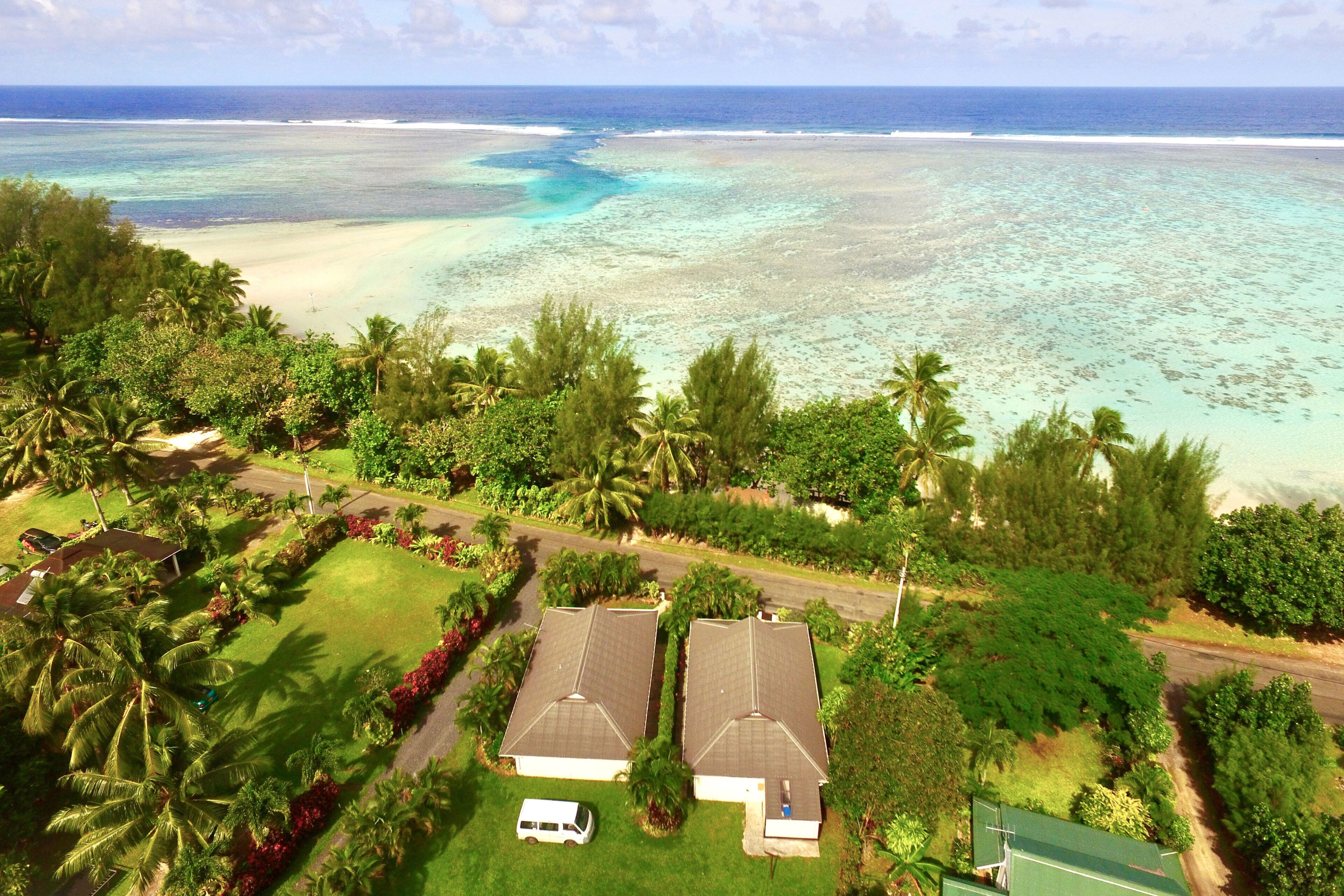 An aerial view of a coastal residential area with two houses, a road, and a beach with clear turquoise waters.