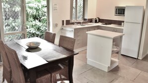 Cuina moderna amb zona de menjador, amb una illa central, armaris blancs i vista a la verdura exterior.