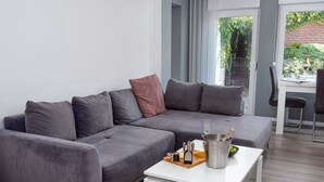 Un salon moderne avec un canapé d’angle gris, une table basse blanche sur laquelle se trouve une bouteille de champagne, et une vue sur un jardin par une porte vitrée.
