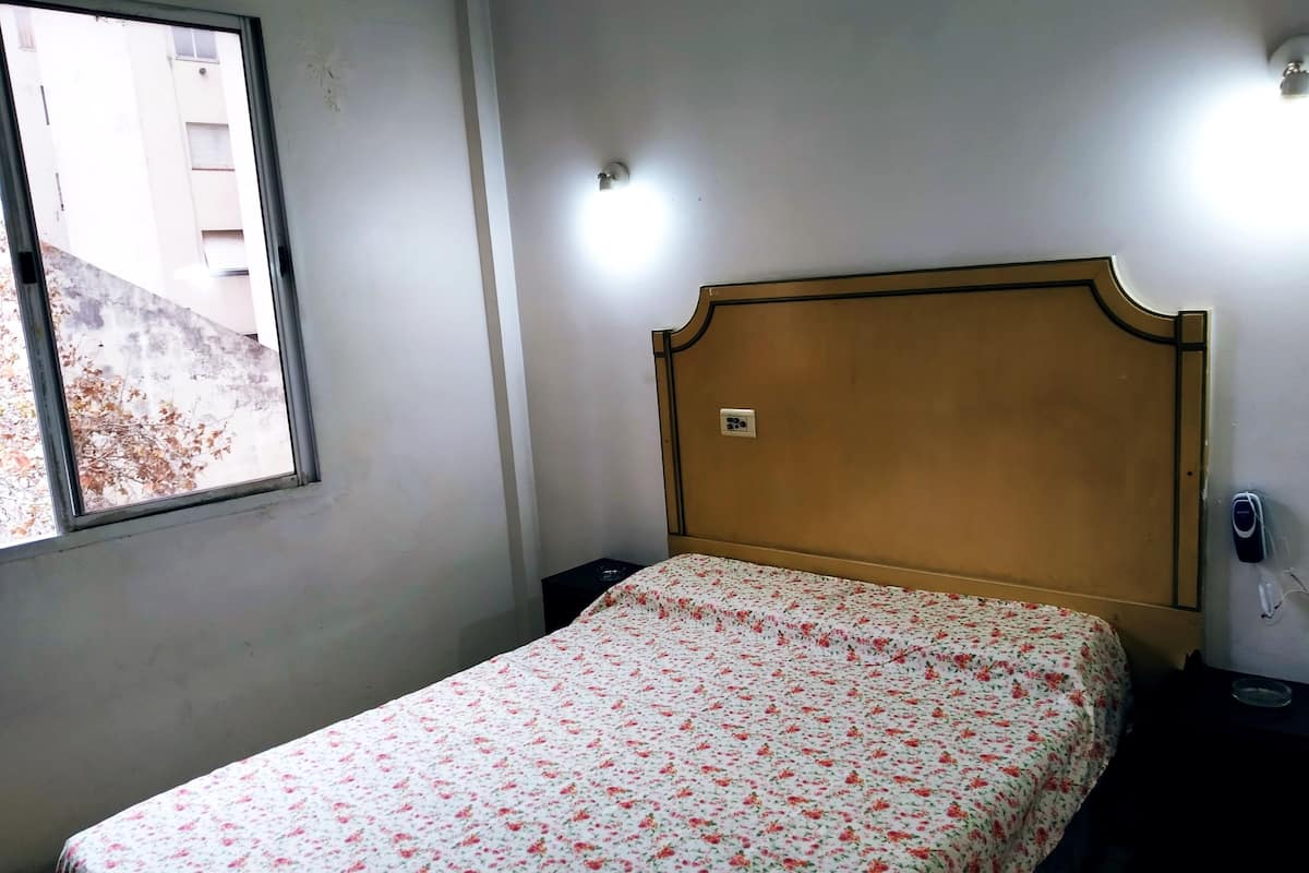 Une petite chambre simple, avec un lit simple, une table de chevet, une lampe de chevet, une fenêtre et un luminaire fixé au mur.