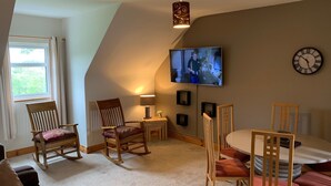 A cozy living room with a sloped ceiling, a television mounted on the wall, a clock, and a window with curtains. - Racecourse Lodge Apartment (Clifden)