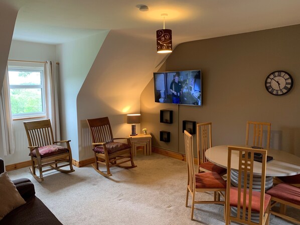 A cozy living room with a sloped ceiling, a television mounted on the wall, a clock, and a window with curtains.