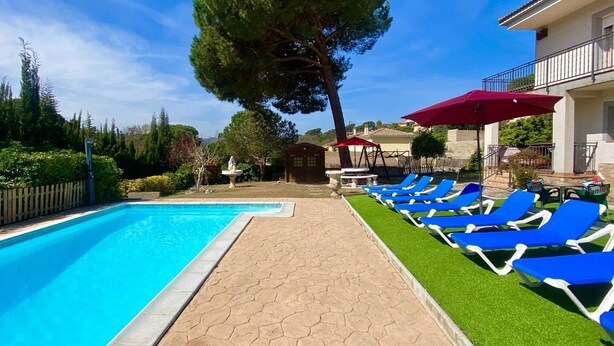 Una piscina con acqua blu, circondata da un percorso lastricato. Sul lato destro si vedono lettini blu e un ombrellone rosso.